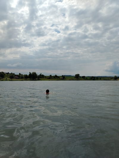 A person is swimming in the middle of a body of water. The person's head and shoulders are visible above the water. The water is calm with ripples. In the background, there is a treeline with some houses and a cloudy sky. The scene is located in Hodyliv, Ukraine.