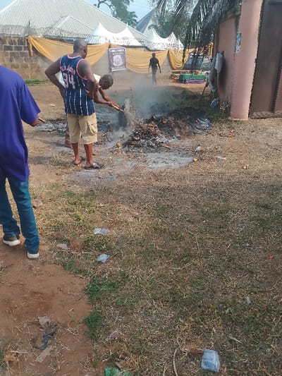 This image captures an outdoor scene in Ufuma, Nigeria. In the central mid-ground, a smoldering pile of dark organic material and debris emits light grey smoke and ash. A male subject, wearing a light-colored short-sleeved top and dark trousers/shorts, is bent over this pile, actively using a stick or similar tool to manipulate its contents. To his immediate left, a second male subject stands upright, wearing a blue and white striped sleeveless jersey with the number "94" on the back and khaki cargo shorts, with dark flip-flops. A third individual, partially visible in the left foreground, wears a purple shirt and blue jeans.

In the background, behind a low cinder block wall topped with barbed wire, a series of white event tents are visible, draped with yellow fabric. A banner or sign featuring text and an image is affixed to the yellow fabric. A fourth, silhouetted individual is visible walking in the distance behind the tents. To the far right, a light-brown colored building with a dark brown door or gate is present. A dark-colored vehicle is parked near the tents, and several green tables or benches are also visible.

The ground consists of dry, unpaved earth mixed with sparse, dry grass. Scattered small pieces of plastic and other debris are present on the ground. The lighting indicates daytime conditions.