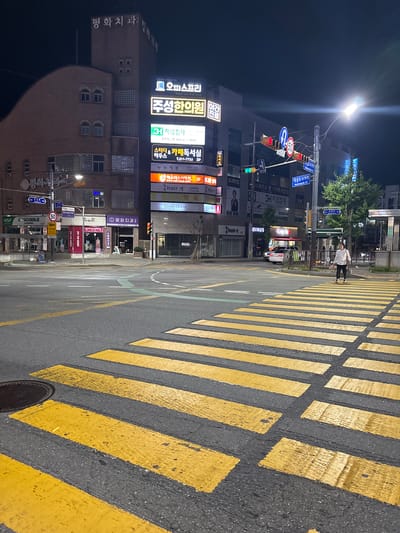 This is a nighttime, eye-level shot of a street scene in Cheongju-si, South Korea. The image is oriented vertically. The foreground is dominated by a pedestrian crossing with bright yellow stripes, indicating a street intersection. To the left, a multi-story building is illuminated by various signs.
The building's facade features several businesses with prominent Korean signage, including "평화치과" (Peace Dental Clinic), "주성한의원" (Juseong Oriental Medicine Clinic), and "누누정원" (Nunu Garden Flower Plant). Other visible businesses include "오피스프리" (Office Free), "슈팅디&카페독서실" (Shooting D & Cafe Study Room), "엑소더스어학원" (Exodus Language Academy), and "Gong cha". Traffic lights are visible, showing a red light in one direction and a green light in another. A street lamp shines brightly from the upper left of the frame.
In the middle ground, two people are crossing the street on the pedestrian walkway, one dressed in a white top and dark pants, the other in dark clothing. A white van is parked nearby. The overall atmosphere is that of a typical urban street at night, with artificial lights illuminating the scene. There is no indication of specific weather conditions, but the darkness suggests it is after sunset.
