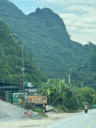 The image depicts a roadside scene in Hà Giang 2 Ward, Vietnam. A large, green, industrial vehicle is parked next to a pile of grey rock and gravel. A yellow dump truck with the number "23U 00475" is parked directly behind it. Adjacent to the truck, there is a pile of cylindrical pipes. Lush vegetation, including banana plants, grows nearby. A road runs along the right side of the frame, with a person riding a motorbike. In the background, a densely vegetated mountain dominates the landscape. Utility poles and power lines are also visible, adding to the industrialized backdrop.