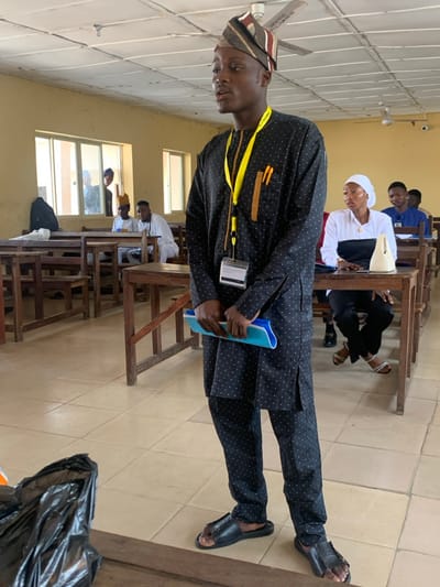 The image depicts an indoor scene within an educational facility, likely a classroom or lecture hall, situated in Abobo, Nigeria, as indicated by "POLYTECHNIC" text on a lanyard.
In the foreground, a young Black male stands, facing slightly right with his mouth partially open, suggesting he is speaking. He wears a black long-sleeved tunic and matching trousers adorned with white polka dots, a striped cap, and black open-toed sandals. A yellow lanyard with an ID holder and two pens is around his neck. He holds a light blue folder or book with both hands at waist level.
The room features light-colored walls, light-brown tiled flooring, and multiple wooden desks with attached benches arranged in rows. Several rectangular windows along the left wall show external light and faint reflections of individuals. Two white ceiling fans are visible overhead.
In the mid-ground, on the right side, a young Black female is seated at a desk, looking towards the standing male. She wears a white top with a black mid-section, black trousers, a white head covering, and light-colored sandals. A light-colored handbag rests on her desk. Behind her, another male in a blue shirt is partially visible. On the left side of the mid-ground, several males are seated at desks; one wears a light-colored traditional outfit and a yellow cap, and a black backpack is visible on a desk near the windows.
A black plastic bag is partially visible on the floor in the bottom left corner of the frame.