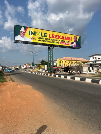 The image shows an outdoor scene in what appears to be Abeere, Nigeria. The main subject is a large billboard promoting the candidacy of "IMOLE LEEKANSI," labeled as "GOVERNOR ADEMOLA JACKSON NURUDEEN ADELEKE FOR 2ND TERM," alongside BARR PELUMI OLAJENGBESI. The billboard is situated beside a road with black and white painted curbs. In the background, there are buildings, including a yellow structure. There is a glimpse of a person standing by the side of the yellow structure with another person by their side and vehicles and street lighting further down the road. The sky is visible with clouds. The image is oriented at an angle, with the road running from the top right corner towards the bottom left.
