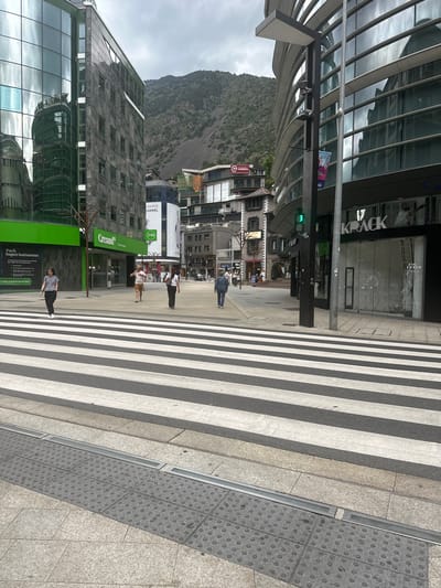 An urban street view in Andorra la Vella, Andorra, captures a bustling commercial area with modern architecture set against a dramatic mountainous backdrop under an overcast sky. A wide zebra crossing dominates the foreground and mid-ground, paved with light grey and black stripes, contrasting with the granite-tiled sidewalks, some featuring textured tactile paving.
Several modern buildings with reflective glass facades and angular designs line the street. A prominent green traffic light is visible on a street pole. Numerous pedestrians are scattered across the scene; some are walking on the crosswalk, while others stand on the sidewalks or near storefronts, engaged in typical daytime city activity.
Visible text on businesses and signs includes "Creand Crèdit Andorrà" on a large green sign, "Pack SuperAutònoms" and "viatges prémium per Europa" also on a green sign. Other visible text includes "KRACK" on a storefront, "CHANCE CHANEL" and "(24h)" on a white building, "MODERNES" (likely "MODERNA") on another building, and "play space" on a shop window. The scene depicts a typical daytime city atmosphere.