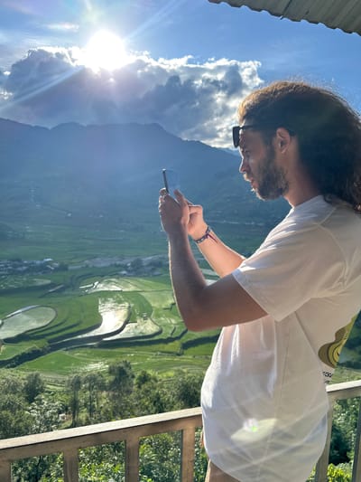 A man with long, dark curly hair and a beard, wearing a white t-shirt and sunglasses perched on his head, stands on a balcony in Tú Lệ, Vietnam. He is viewed in profile, facing right, and is holding a dark-colored smartphone with both hands, appearing to photograph the panoramic landscape. A blue and purple woven bracelet with a white eye-like bead is visible on his left wrist. The foreground features a light-colored balcony railing and the edge of a dark roof overhead. The background reveals a vast mountainous valley filled with vibrant green terraced rice fields, many reflecting the bright sunlight from their water-filled sections. Distant mountains rise under a bright blue sky with scattered clouds. The sun is intensely bright, positioned high in the sky to the left, casting strong light and creating lens flares. Small clusters of buildings are visible within the valley amidst the terraced fields. Lush green trees cover the slopes below the cultivated areas.
