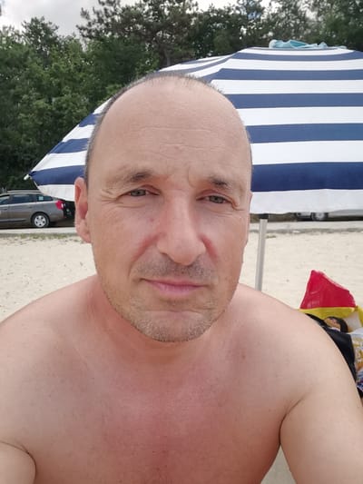 A selfie taken on a beach in Osenovo, Bulgaria, shows a shirtless middle-aged man with thinning hair, wearing a slight smile. He is positioned in the foreground, with a large blue and white striped beach umbrella behind him, providing shade. The background reveals a sandy beach, a line of trees, and a dark-colored vehicle parked near the water. To the right, a red and yellow bag and other beach items are visible. The man's skin appears slightly flushed, indicating sun exposure.