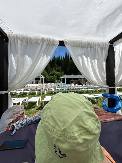 The image shows a personal perspective from under a canopy, looking out at an outdoor swimming pool area. In the foreground, a person wearing a light green bucket hat is seated on a dark surface, with a smartphone, a patterned fabric item, and a blue inflatable ring visible. The canopy above is white and draped with white sheer curtains. Beyond the canopy, rows of empty white lounge chairs are arranged on a grassy area. In the background, a pool with several people swimming is visible, along with trees and a clear blue sky. The location is identified as Nechkino, Russia.