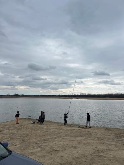 The image displays a daylight scene of a group of six individuals on a sandy shoreline next to a large body of water in Любен, Bulgaria. The sky is uniformly overcast with numerous grey clouds.
A dark blue vehicle's hood and windshield are partially visible in the bottom-left foreground, parked on the dirt bank. Five individuals are positioned along the water's edge. From left to right: one person in light-colored pants and a dark jacket stands near the water; a group of three individuals, including two in dark jackets, are clustered near a white folding chair and a blue bucket on the sand; one person holds a tall, black fishing rod vertically upright, extending high into the frame; and a final person in dark clothing stands to the right, facing the water.
The body of water occupies the middle ground, exhibiting light ripples on its surface. Across the water, a distant, low horizon line features a strip of dark, bare trees or vegetation. Multiple fishing rods are present on the bank or held by the individuals.