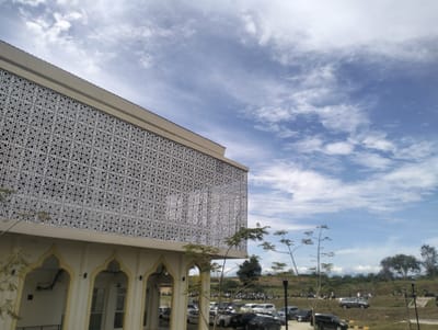 The image depicts a modern building with distinctive patterned facade in Reuleut Timu, Indonesia. The building features a cream-colored base with arched openings and an upper level clad in a white, intricate geometric screen. The sky above is a vibrant blue with scattered white clouds, suggesting a bright, daytime scene. In the foreground, a parking lot is visible with several cars and motorcycles parked. A few sparse trees are scattered across the scene, with some foliage on the left side of the building and more distant trees forming a treeline on the horizon. The overall environment appears to be an open area, possibly a public space or educational institution, given the modern architectural style and the presence of a parking area.