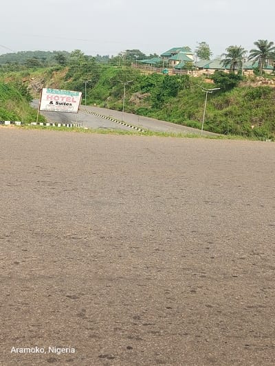The image shows a paved road with a sign for the "Mamu Royal Hotel & Suites" visible in the foreground. The road is empty, but there are two street lights on the road and two more on the grassy hilltop. The road leads up to a hilly area, with a dense growth of green vegetation and a few buildings in the background. There is a white sign with black lettering with Arabic writing on it located at the top of the hill. The location is a road in Iyin, Nigeria, at coordinates 7.661 latitude and 5.166 longitude. The sky is clear, with no clouds visible. The image appears to have been taken during the day, possibly in the early afternoon. The photograph has a shallow depth of field, with the sign in the foreground in sharp focus and the background slightly out of focus.