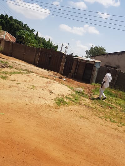 In Palade, Nigeria, a person dressed in white walks along a dirt path beside a row of simple buildings. The buildings appear to be made of concrete with corrugated metal roofs and feature open doorways with barred windows. Vegetation, including trees and grass, grows along the path and behind the buildings. Overhead, a few wispy clouds drift across a bright blue sky, suggesting it is daytime and the weather is clear. The scene has a rustic, everyday feel, with no indication of a specific event or strong emotions. The focus is on the mundane, capturing a moment of solitary movement through a common environment.