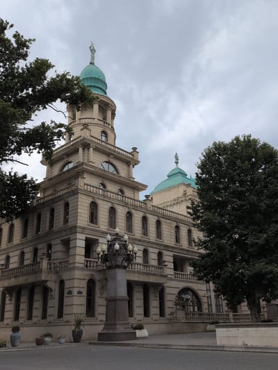 The image shows the exterior of a grand, light-colored stone building with an ornate architectural style. The building features multiple levels with arched windows and balconies adorned with decorative railings. A prominent feature is a turquoise-domed tower, topped with a statue or spire, and another similar dome is visible on a lower section of the building. Green trees with dense foliage partially obscure the view of the building.

The perspective is from a low angle, looking upwards towards the building and the sky. The sky is overcast and cloudy, suggesting a daytime scene, likely morning or afternoon. The building appears to be situated alongside a street or road, with a grey asphalt surface visible on the right side of the image. Several potted plants are placed near the base of the building along the sidewalk, including one large blue vase. A decorative street lamp with multiple globes is also visible, attached to a stone column.

There is some visible text on a sign above an arched entrance towards the lower right of the building; the word "KASS" is clearly legible. The overall impression is one of historical grandeur and official architecture, potentially a government building, museum, or historical landmark in Ganja, Azerbaijan. No people or specific activities are evident in the image.