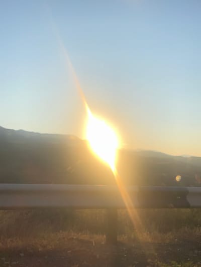 The image depicts a bright sunset over the rolling hills surrounding Arpi, Armenia. The sun, a brilliant orb of light, is positioned in the upper center of the frame, casting a warm glow across the sky. Lens flares and light rays emanate from the sun, creating a hazy and ethereal effect. In the foreground, a guardrail runs horizontally across the image, separating the viewer from the landscape beyond. The ground beneath the guardrail is covered in dry grass and scattered stones. The distant hills are silhouetted against the vibrant sky, their contours softened by the sunset light. The overall impression is one of serene natural beauty, characteristic of the Armenian countryside at dusk.