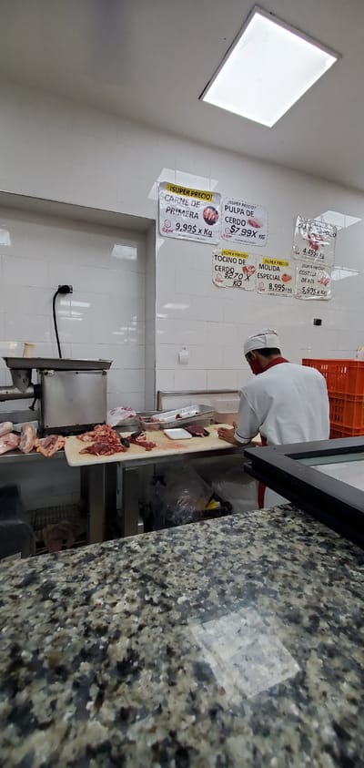 The image captures the interior of a butcher shop or meat department, likely in Barquisimeto, Venezuela, during daylight hours, illuminated by bright artificial lighting. A male butcher, wearing a white uniform and a red face mask, is actively preparing meat behind a counter. He stands over a large wooden cutting board, where various cuts of raw meat are visible, some already sliced. A meat grinder is partially seen to his left, and additional raw chicken pieces are stacked below the counter. The work area features white tiled walls, with a bright square ceiling light visible in the upper left. A black electrical cord is plugged into an outlet on the wall. The preparation area is separated from the viewer by a speckled dark granite counter.

Several laminated price signs are affixed to the tiled wall, advertising "super prices" for different meat products:
*   "¡SUPER PRECIO! CARNE DE PRIMERA" (First-class Meat) is priced at "9,99$ x Kg".
*   "¡SUPER PRECIO! PULPA DE CERDO" (Pork Pulp) is "$7.99X KG".
*   "¡SUPER PRECIO! TOCINO DE CERDO" (Pork Belly/Bacon) is priced at "$2.70 X KG".
*   "¡SUPER PRECIO! MOLIDA ESPECIAL" (Special Ground Meat) is "8,99$ x KG".
*   "¡SUPER PRECIO! COSTILLA DE RES" (Beef Ribs) is "5,99$ x KG".
*   "CRAYON DE POLLO" (Chicken Thigh/Drumstick) is listed at "4.49$ Kg".

The scene suggests a clean, well-organized commercial environment focused on meat preparation and sales.