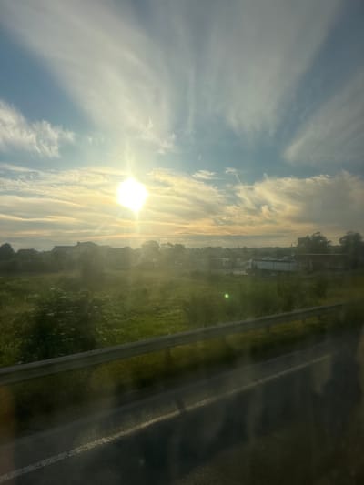 The image depicts a rural landscape at sunrise or sunset, characterized by a bright sun and wispy clouds. In the foreground, a metal guardrail is visible, casting shadows on a paved surface that appears to be a road. Beyond the guardrail, a grassy field stretches out, with scattered green vegetation. In the middle ground, several buildings are discernible, including what appears to be a large residential or commercial structure, along with other smaller buildings and trees. Some trucks and what might be industrial buildings are also present in the distance, suggesting a rural or semi-rural area on the outskirts of a settlement. The overall atmosphere is serene, with the warm light of the sun illuminating the scene and creating a slightly hazy effect. The location is identified as Kamianopil, Ukraine.