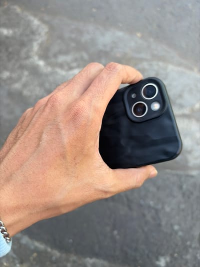 A person's hand is holding a black iPhone 13 Pro in Pekalongan, Indonesia. The phone's camera module, featuring three lenses, is visible. The hand is positioned over a textured grey surface, possibly a sidewalk or pavement. The lighting suggests it is daytime.