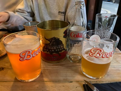 A table arrangement features two beer glasses with "Beer" emblazoned on them, alongside a tin of "TEMERITY JONES LONDON PIMENTON DE LA VERA," a glass bottle of Diet Coke, and two additional glasses. The setting seems to be indoors, with a person in an "athletic" sweatshirt visible in the background. The scene is set on a wooden surface.