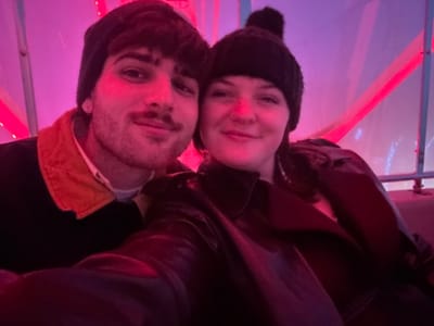 A selfie captures two people indoors. The scene is lit with pink and red light, creating a vibrant atmosphere. One person wears a black beanie with a pom-pom and a dark jacket. The other person sports a beanie and a black top with an orange collar. Their faces are close, suggesting a close relationship. The background is slightly blurred and features translucent panels.