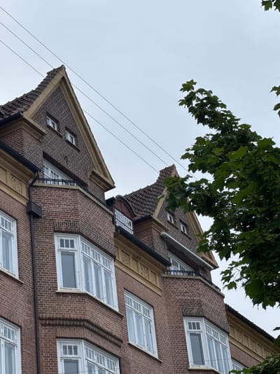 This image shows a portion of a red brick apartment building in Frederiksberg, Denmark. The building features ornate yellow trim around the gables and upper facade. Several bay windows with white frames are visible, some with multiple panes. The roof is tiled, and a section of the building has a rounded corner. Overhead, utility wires are strung across the sky, which is overcast. A large tree with green leaves partially obscures the right side of the building. The scene is a street-level view looking upwards at the building's facade.