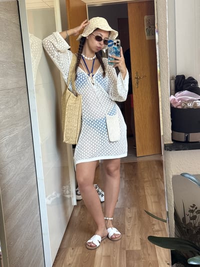 A woman stands indoors, taking a mirror selfie. She is wearing a white crochet cover-up dress over a blue bikini, a straw hat, sunglasses, and white slide sandals. She accessorizes with a shell necklace and a straw tote bag. The background includes a closet door, a wooden door, and a shelf with clothing and a plant. The floor is made of wood-look laminate. The scene appears to be in a domestic setting.
