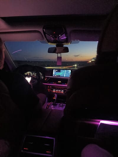 This image captures the interior of a vehicle at night or late dusk in Troyanovo, Bulgaria. The car's cabin is illuminated with prominent purple/pink ambient lighting along the dashboard, center console, overhead console, door panels, and rear passenger area.
An adult male driver is positioned on the left, viewed from behind. He holds an illuminated smartphone in his left hand while the vehicle is in motion. The digital instrument cluster behind the steering wheel displays a speedometer indicating approximately 50-60 units and a time of 21:44. The central infotainment screen shows a navigation map with a green route. Below it, the climate control panel indicates a temperature setting of 22.0°. The automatic gear selector and various illuminated controls are visible on the center console. A rearview mirror hangs from the ceiling, with a suspended air freshener or charm.
A front passenger is seated on the right, with only the back of their head and upper torso visible, largely obscured by shadow. The lower legs of a rear passenger are visible in the bottom right foreground, illuminated by the interior's purple lighting.
Through the windshield, the twilight sky transitions from dark blue at the top to an orange-yellow gradient at the horizon. Distant lights, indicative of other vehicles or infrastructure, are visible on what appears to be a road or highway. A distinct purple light streak is visible in the upper left portion of the windshield.