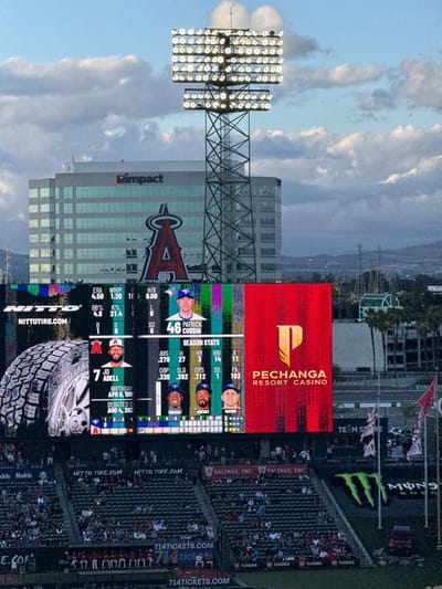 The image captures a vibrant scene at a baseball stadium, likely Angel Stadium in Anaheim, United States, under a cloudy, late afternoon or early evening sky. The focal point is a massive video scoreboard displaying game information and advertisements, backed by a large building with the "impact" logo and a prominent red Angels logo. A tall stadium light tower stands in front of the building, illuminating the field.

The scoreboard features player statistics, showing "46 PATRICK CORBIN" (a pitcher, possibly for the Toronto Blue Jays given the logo) with ERA, WHIP, H/9, BB%, and K% stats. Below, "JO ADELL" (an Angels player, #7) is highlighted with his birthday, debut date, and season stats including AVG, H, HR, RBI, R, OBP, SLG, OPS, SB, and PA. Images of other players like "SANCHEZ," "JIMENEZ," and "VARSHO" are also visible. On the right side of the scoreboard is a large red section with the "PECHANGA RESORT CASINO" logo.

Below the scoreboard, sections of stadium seating are visible, filled with numerous spectators. Various advertisements and logos are plastered throughout the stadium, including "NITTO TIRE.COM," "SALINAS, INC.," "714TICKETS.COM," "Modelo," "TUNDRA," and a large "MONSTER" (Monster Energy) logo. The overall setting is that of an active professional baseball game, with bright lights and digital displays creating an energetic atmosphere despite the cloudy conditions.