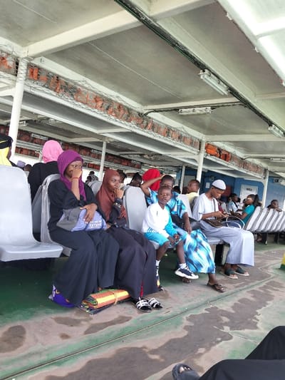 The image depicts the interior of a passenger ferry or vessel, likely operating near Mkomani ward, Kenya. Approximately twelve individuals are visible, seated in rows of grey, molded plastic seats. The setting is a covered deck with a light-colored ceiling supported by structural beams, from which fluorescent tube lights are suspended. Orange life jackets are stowed in mesh netting racks above the seating areas on both sides of the vessel. The floor is dark green, showing signs of wear and moisture.
In the foreground, a woman wearing a black abaya and a purple headscarf sits on a grey seat, holding a blue and grey shoulder bag. Her right hand is near her mouth. Next to her, a second woman in a black abaya and a brown headscarf is also seated, with her hand near her chin. Below their seats, a rolled, multi-colored mat is placed on the floor, secured with a yellow strap.
To the right, a child wearing a white shirt and blue shorts sits on a grey seat, looking towards the camera. An adult, partially obscured, sits next to the child, wearing a blue and white patterned garment. Further right, a man with a white kufi (cap) and a light-colored thobe (garment) sits, looking down at a mobile phone in his hands. A dark bag is on his lap. Other passengers, some wearing head coverings, are visible in the background, seated or partially obscured. The overall scene suggests transit on a public ferry.