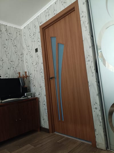 The image depicts an interior scene, likely a hallway or room, with a wooden door as the central element. The door is made of a light brown wood veneer with two vertical, narrow, blue glass insets. A silver door handle is visible on the right side of the door. To the left of the door, a wall is covered in light-colored floral wallpaper with a repeating pattern of grey stems and flowers. A dark wooden cabinet with two doors is situated against this wall, topped with a black computer monitor, a keyboard, and a decorative wooden object with three candle holders. A small digital clock with a white face is also on the cabinet. To the right of the wooden door, a sliver of a different door or wall is visible, appearing to be made of a reflective material, possibly glass, with a circular pattern. The floor is covered with laminate flooring that mimics dark wood planks. The scene appears to be located in Zhovti Vody, Ukraine.
