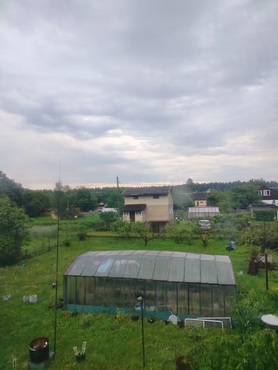 The image depicts a rural outdoor scene, possibly in the Дзешчанскі сельскі Савет, Belarus, under an overcast sky. In the foreground, a large, rectangular greenhouse with a green frame and transparent glass panels is situated on a grassy area. To the left of the greenhouse, there's a dark cylindrical object on a base, possibly a rain barrel. Several white bags are scattered around the immediate vicinity of the greenhouse. A tall, thin pole, likely an antenna or support, stands to the left of the greenhouse.
Further back, a two-story house with light-colored siding and a dark roof is visible, surrounded by green trees and vegetation. A smaller pink building and another house with a dark roof and white trim are also present in the mid-ground. In the background, a dense line of trees forms the horizon. The overall environment is characterized by lush green grass, various trees, and a slightly neglected, rural aesthetic. There are no visible people in the image, but various gardening tools and structures suggest human presence and activity. The lighting is subdued due to the cloudy weather.
