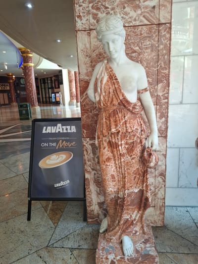 The image depicts an indoor scene with a marble statue of a woman on the right, adorned in a draped garment. To the left of the statue, an A-frame sign displays a Lavazza coffee advertisement. The sign features a close-up of a coffee cup with latte art, the Lavazza logo, and the tagline "ITALIAN COFFEENESS ON THE Move". The statue stands against a pillar made of patterned marble. In the background, a walkway with tiled flooring is visible, along with other architectural elements such as columns and lighting fixtures. The location appears to be a public indoor space, possibly a shopping center or a hotel lobby, within Trafford, United Kingdom, given the context provided.