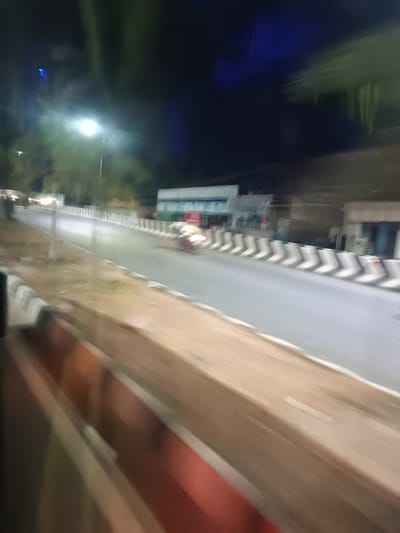 The image depicts a blurred, nighttime street scene in Anantapur, India, captured from a moving vehicle. The lower foreground shows an out-of-focus, reddish-brown horizontal structure, likely part of the vehicle from which the photograph was taken.

The central feature is a multi-lane paved road, illuminated by overhead streetlights. A median or lane divider runs down the center of the visible road, constructed from a series of concrete barriers, each painted with alternating black and white diagonal stripes. To the left of these barriers, a dirt or gravel shoulder is present.

A single vehicle, appearing to be a motorcycle or scooter, is visible on the road, exhibiting significant motion blur. Its headlights or taillights are illuminated, appearing as a bright, streaked white light.

In the background on the right, several indistinct commercial buildings with illuminated signs are visible, one of which has a light blue and white facade. On the left, beyond the road and a prominent streetlight on a pole, sparse vegetation, possibly trees, can be discerned against the dark night sky. No individuals are clearly visible within the scene.