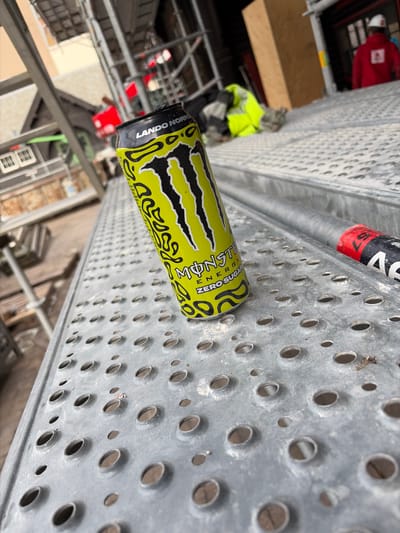 A skewed, high-angle shot depicts a perforated metal platform, likely part of scaffolding, with two partially visible energy drink cans – one Monster Energy Zero Sugar in green and black, and another in red and black. A construction worker in a yellow safety vest is visible in the background. The location is Ostend, Belgium, as evidenced by the "LANDO NORRIS" text on the can, a collaboration with the Formula 1 driver.