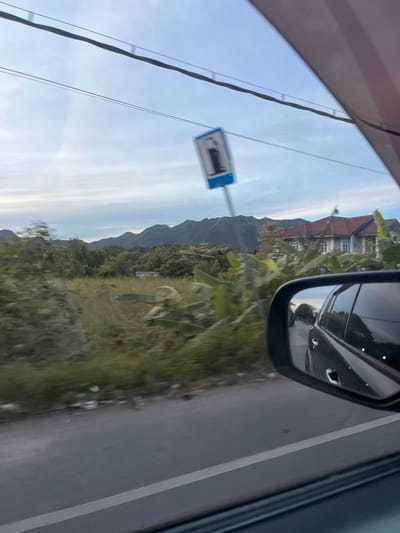 The image captures a view from a moving vehicle in Beuradeun, Indonesia. In the foreground, the side mirror of the car shows a reflection of the road behind and the vehicle itself. The road is paved and appears to have a white line marking the lane. To the left of the road, there is an expanse of dry grass and some scattered vegetation. In the distance, a large house with a red tiled roof is visible, surrounded by greenery and trees. Further beyond, a range of lush green mountains forms the backdrop under a cloudy sky. A blue sign with a white symbol, possibly indicating a facility or restriction, is visible on a pole in the middle ground. The overall scene depicts a rural or semi-rural environment, characteristic of the outskirts of a town or city.