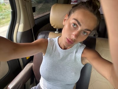 A single adult female is depicted in a close-up selfie taken inside a vehicle in Paje, Tanzania. She possesses light skin, green eyes, and her dark hair is styled in an updo, likely a bun. Her facial expression is neutral, and she directs her gaze forward into the camera. She is attired in a light grey ribbed sleeveless top. The vehicle's interior features a passenger seat with a tan headrest and upper backrest, transitioning to a dark brown or grey lower and side upholstery. A beige seatbelt is visible on the left side of the frame. Through the vehicle's window, a blurred outdoor environment comprising green vegetation and a dirt road is discernible, indicating a daytime, possibly rural or semi-rural, setting.