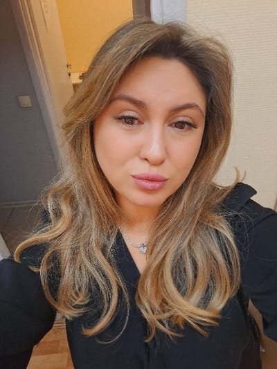 A female subject is centered in a close-up, frontal portrait, occupying the majority of the frame. She has fair skin, light brown eyes, and medium-length wavy hair with blonde highlights. Her lips are lightly colored with pink lipstick and appear slightly pursed. She is wearing a black garment, likely a shirt or blouse, and a silver-colored necklace with a heart-shaped pendant is visible at the base of her neck. The background is out of focus. To the subject's left, a light gray wall or door frame is visible with a white rectangular switch or outlet, and beyond it, a glimpse of a light yellow wall and a white fixture suggests an adjacent room. To the subject's right, a light beige textured wall is visible. The scene is indoors, under artificial lighting. The specific location within the city of Teykovo, Russia, cannot be determined from the visual evidence in the image.