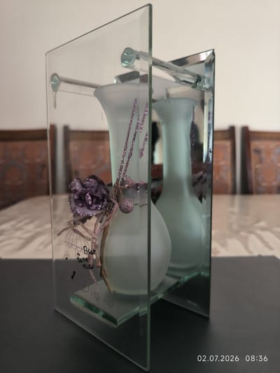 A glass decorative object with two frosted vases stands on a dark surface. The object has a frame structure made of glass, with a horizontal rod supporting the top. The vases are decorated with purple glittery elements and a dried flower. Text, including "Bear," "fame," and "nia," is written on the glass. In the background, out of focus, are wooden chairs and a table with a patterned tablecloth. The timestamp "02.07.2026 08:36" is visible in the lower right corner. The scene is indoors. There is no indication of the city of Kattasu, Uzbekistan.