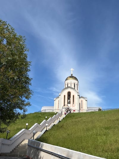 A majestic Eastern Orthodox church stands under a brilliant blue sky, prominently featuring a large, gleaming golden dome topped with a golden Orthodox cross. The church, with its light-colored facade, arched windows, and intricate architectural details, is situated on a verdant hillside covered in lush green grass.
A long, zigzagging concrete staircase with white steps and gray railings ascends the slope towards the church. Spherical light fixtures are regularly spaced along the staircase, suggesting its use after dark. A dark metal fence runs along the top of the grassy hill near the church building. In the lower-left foreground, a tree with dense green foliage partially frames the scene.
The sky is bright and clear with only a few wispy white clouds, indicating a sunny day, likely morning or afternoon, given the strong illumination on the church. One person wearing a red top is visible near the base of the church, adjacent to the staircase. The architectural style and prominent cross are characteristic of Eastern Orthodox churches, aligning with the location context of Varvarovka, Russia. The scene conveys a sense of grandeur and peacefulness. No visible text is present in the image.