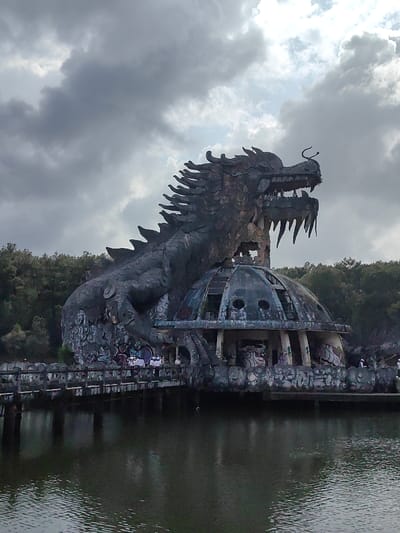 This image depicts the abandoned Ho Thuy Tien water park structure in Kim Long Ward, Hue, Vietnam. A substantial, dark grey, weathered dragon sculpture is centrally featured, its scaled body encompassing a dilapidated, dome-shaped concrete building. The dragon exhibits an open mouth with visible teeth, prominent dorsal spines, and horn-like protrusions on its snout. The dome structure beneath it is partially open at the top, features arched windows, and displays extensive graffiti on its lower, columned section. A concrete bridge with railings extends from the left foreground across dark, still water, connecting to the base of the dome. Approximately five to seven individuals, clad in casual attire including some white shirts, are observed standing on the bridge and near the structure's base, appearing to explore the site. The background consists of dense green trees. The sky is predominantly overcast with dark grey clouds, with a segment of brighter, partially sunlit clouds visible on the upper right, illuminating the scene from that direction. The entire structure shows advanced signs of weathering and decay.