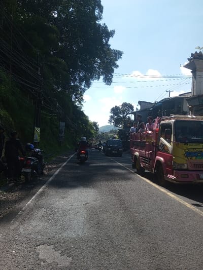 This image depicts a daytime street scene in Ban Phonhxavanh, Laos, characterized by a paved asphalt road, roadside vegetation, and various vehicles and people.

The foreground features a two-lane asphalt road exhibiting patches of repair. On the immediate left, a steep, densely vegetated embankment rises, featuring large trees and multiple utility poles with a complex network of overhead wires. Two rectangular banners are affixed to these poles. At the base of the embankment, near the left shoulder, two individuals in dark clothing stand beside two parked motorcycles. One motorcycle, dark in color, displays a partially visible Lao license plate.

On the right side of the road, a large, brightly colored pink and yellow truck, modified with an open cargo bed, is positioned, facing away from the viewer. The cargo bed contains at least seven passengers. Behind the truck, a building with an ornate, multi-tiered rooftop structure is partially visible, alongside other buildings and trees further down the road.

Mid-ground shows a single individual wearing a white helmet, riding a red scooter or motorcycle away from the viewer on the left side of the visible lane. Further down the road, a dark minivan or SUV is visible, along with other distant vehicles.

The sky above is clear blue with scattered white cumulus clouds. Strong sunlight is evident, casting shadows across the road and from the trees. Hills are visible in the distant background.