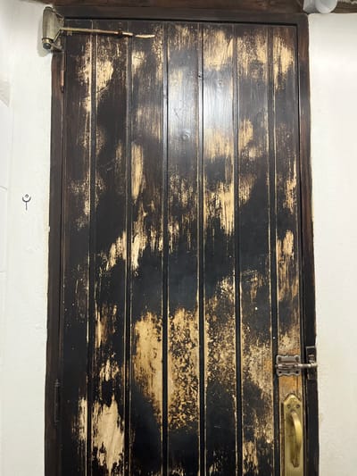 A close-up of a dark wooden door with a brass doorknob and latch. The door is made of vertical planks, and the paint is chipped and peeling, revealing the lighter wood underneath. There is a metal doorstop on the left side of the door, as well as a small symbol drawn on the white wall adjacent. The scene is in the city of Agüimes, Spain, located at the coordinates 27.907 latitude and -15.447 longitude.