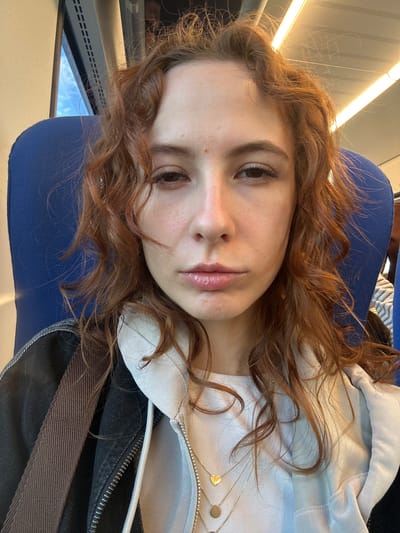 A young woman with reddish-brown, curly hair is lying on a blue seat, likely on a train or bus. She is fair-skinned with pink lips and is wearing a light-colored hooded sweatshirt with a zipper and a dark jacket. She has a gold necklace with small charms. Outside the window, there are glimpses of blue sky and some structures that can be seen due to the window.
There is nothing to imply the exact location within Stara Pazova Municipality, Serbia.