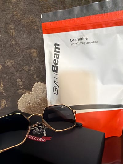 The image shows a GymBeam L-carnitine package (net weight: 250 g Lemon-lime) next to a pair of gold-rimmed sunglasses and a black case with "CULLINO" printed on it. The items are placed on a surface with a floral doily. The location is unknown; no features indicate it is Brusen, Bulgaria.