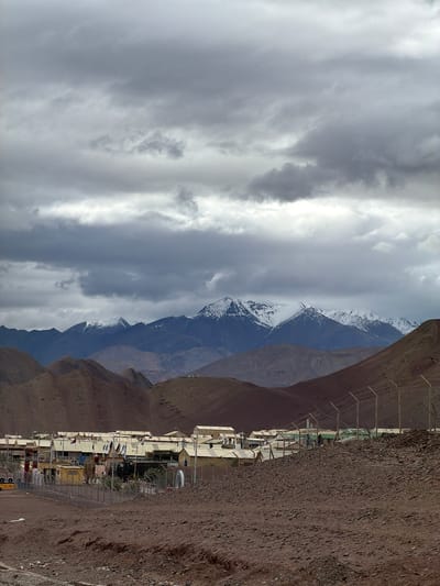 In Nyoma, India, an overcast sky looms over a mountainous, arid landscape. In the middle ground, a settlement of beige, prefabricated buildings with light-colored roofs forms a compound. A wire fence encloses the settlement, with several flags visible, some atop buildings and others along the fence line. A vehicle, possibly a truck, is parked within the compound. In the foreground, the ground is covered in reddish-brown dirt and small rocks. The background is dominated by rolling, barren hills that lead to a distant mountain range with snow-capped peaks.