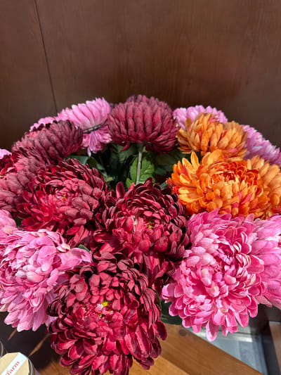 A close-up, overhead view showcases a vibrant bouquet of artificial chrysanthemums. The flowers are arranged in a dense cluster, with their layered petals creating a textured, almost spherical appearance. The color palette is rich and diverse, featuring shades of deep red, fuchsia, bright pink, and a striking orange. The background consists of a dark, warm-toned wooden surface. A portion of a small, cylindrical object with a light-colored label is visible in the bottom left corner. The lighting suggests it is daytime, casting subtle shadows that highlight the texture of the petals. The scene is devoid of people and any specific environmental context suggesting a location within Phu Thuy, Vietnam, or any other city.