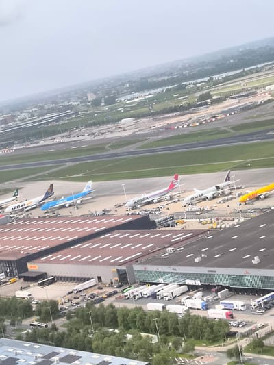 The image presents an aerial view of an airport cargo area, revealing several parked cargo airplanes representing different airlines, including KLM Cargo, Atlas Air, Cargolux, Qatar Airways, and DHL, with a long runway behind them. In the foreground, large cargo warehouses are visible, loaded with trucks at numerous loading bays, suggesting an active freight operation. Trees surround these structures. In the distance, there's a hazy cityscape dotted with water bodies and buildings. Given the airlines and infrastructure present, the image depicts a significant airport cargo hub, potentially in Fischamend, Austria. The environment seems overcast, contributing to the diffused lighting across the scene.
