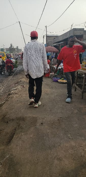 The image depicts an unpaved street scene in Buzove, Democratic Republic of the Congo, likely a market area, under an overcast or hazy sky. The ground is composed of uneven dirt with visible ruts and scattered debris. Overhead, multiple black electrical or communication wires are strung between wooden poles.

In the foreground, two male individuals are walking. On the left, a man with his back to the viewer wears a red baseball cap, a white long-sleeved shirt with a textured pattern, black pants, and white-and-brown sneakers. He is walking away from the camera. On the right, a second man faces forward, slightly turned to his left. He wears a red short-sleeved t-shirt with a yellow McDonald's logo, dark grey jeans, and light blue or grey sandals. His right hand is raised to his forehead, partially obscuring his face.

To the left of the foreground figures, multiple motorcycles are visible on the dirt road. One individual in a red outfit is seated on a red motorcycle. A yellow vehicle is partially visible further in the background on the left. On the right side of the street, rudimentary market stalls are present. One stall has an orange umbrella for shade. Merchandise, including bowls of food items and bottles of liquid, is displayed on wooden tables and on the ground. Buildings constructed from concrete blocks and corrugated metal are visible in the background on the right. Numerous other individuals are visible in the midground and background, populating the street and market area.