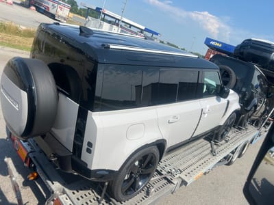 This media file captures a bright, sunny day in St. Marienkirchen bei Schärding, Austria, with a clear blue sky and scattered clouds. The main subjects are two Land Rover Defender vehicles, one white and one black, loaded onto a car carrier trailer.
The white Defender is in the foreground, viewed from its rear-left side. It features a black roof, black wheel arches, black tinted windows, and a distinctive rear-mounted spare tire with a silver cover. The vehicle's license plate, "AA 400 YL," is visible, along with a "90" sticker on the trailer. The trailer itself is made of grey metal with a perforated surface and is secured with straps.
Behind the white Defender, the black Defender is partially visible, also loaded onto the trailer. It has a prominent front grille and headlights. Another vehicle, possibly a trailer or another car, is stacked above it, with a blue sign labeled "AS24" clearly visible.
In the background, a logistics company's truck with the text "H.Z. Logistics" is parked. A fuel station with a sign "International Diesel Service" is also present. The overall scene suggests a moment of transport and logistics, likely related to vehicle delivery or distribution. The lighting indicates it is daytime, possibly mid-morning or early afternoon. The absence of people suggests the focus is solely on the vehicles and their transport.