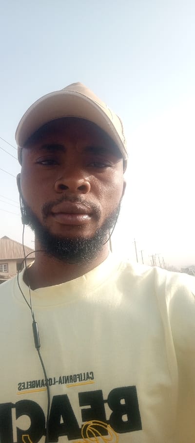 The image displays an adult male captured in a selfie outdoors in Iddah, Nigeria. The individual has a dark complexion and a short dark beard. He is wearing a light-colored baseball cap and a light yellow long-sleeved shirt. The shirt features reversed black and yellow embroidery on the chest, partially visible, consistent with "BEACH CALIFORNIA LOS ANGELES". Black wired earphones with an integrated microphone are visible, draped around his neck and entering his ears. The background on the left side reveals a building with a light-colored corrugated metal roof and light-colored walls featuring white-framed windows. Overhead utility wires are also visible. The right side of the background is largely overexposed and hazy, showing faint outlines of multiple utility poles in the distance under a bright, clear sky.