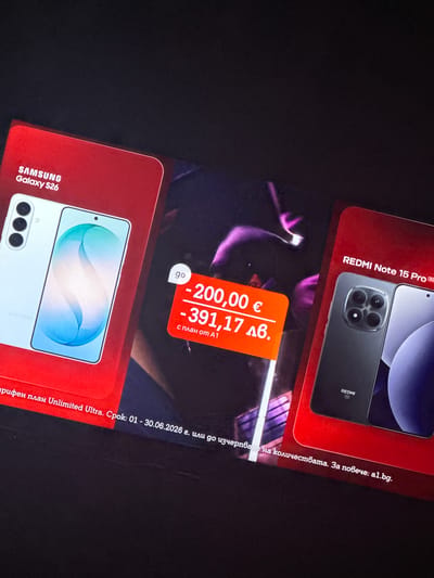 The image shows a promotional display for mobile phones, likely in an advertisement or in-store display. There are two prominent phone models featured: a SAMSUNG Galaxy S26 and a REDMI Note 15 Pro. Both phones are presented on red background cards with their respective brand names and model numbers clearly visible.
In the center of the image, an orange price tag is superimposed over a blurred background image featuring abstract purple lighting and possibly silhouettes of people. The price tag displays two prices: "- 200,00 €" and "- 391,17 лв.", indicating a discount in Euros and Bulgarian Lev. Below these prices, it reads "с план от A1", suggesting these prices are associated with a plan from the provider A1.
The bottom of the image contains text in Bulgarian. The visible portion reads "тариферен план Unlimited Ultra. Срок: 01 - 30.06.2026 г. или до изчерпване на количествата. За повече: a1.bg." This translates to "tariff plan Unlimited Ultra. Period: 01 - 30.06.2026 or while stocks last. For more: a1.bg."
The image appears to be a photograph taken from a slightly elevated angle, with the display elements angled towards the viewer. The lighting in the image is somewhat dim, with the bright colors of the promotional materials standing out against the dark background. There is no visible indication of the city of Smyadovo, Bulgaria, within the image itself.