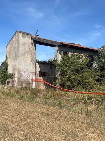 The image shows an abandoned, partially constructed building in Castiglione delle Stiviere, Italy. The building is made of concrete with exposed brickwork in some areas. The roof appears to be damaged, with some tiles missing and wooden beams exposed. Overgrown vegetation, including ivy and trees, covers parts of the structure and the surrounding area.

The scene is enclosed by a wire fence with an orange safety net attached. The ground is covered in dry, matted grass, suggesting a warm, possibly dry season. The sky is a clear, bright blue with a few wispy clouds, indicating it is daytime, likely late morning or early afternoon given the strong sunlight. The overall impression is one of neglect and decay, with nature reclaiming the abandoned structure. There are no people visible in the image. No text is visible.