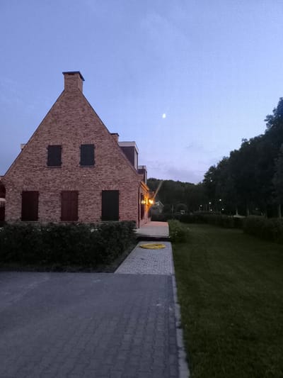 The image captures a twilight scene in Almere, Netherlands, focusing on a two-story brick house with dark shutters on its windows. A prominent chimney rises from the gable roof. A paved walkway leads from a driveway towards the entrance of the house, where outdoor lights are illuminated. To the right of the house, a manicured lawn stretches out, bordered by a dense hedge and a line of trees in the distance. The sky is a dusky blue with a faint moon visible. The environment suggests a residential area, likely a suburban or rural setting within Almere. There are no people or animals visible in the frame, and no overt actions or interactions are depicted. The scene is static and composed.